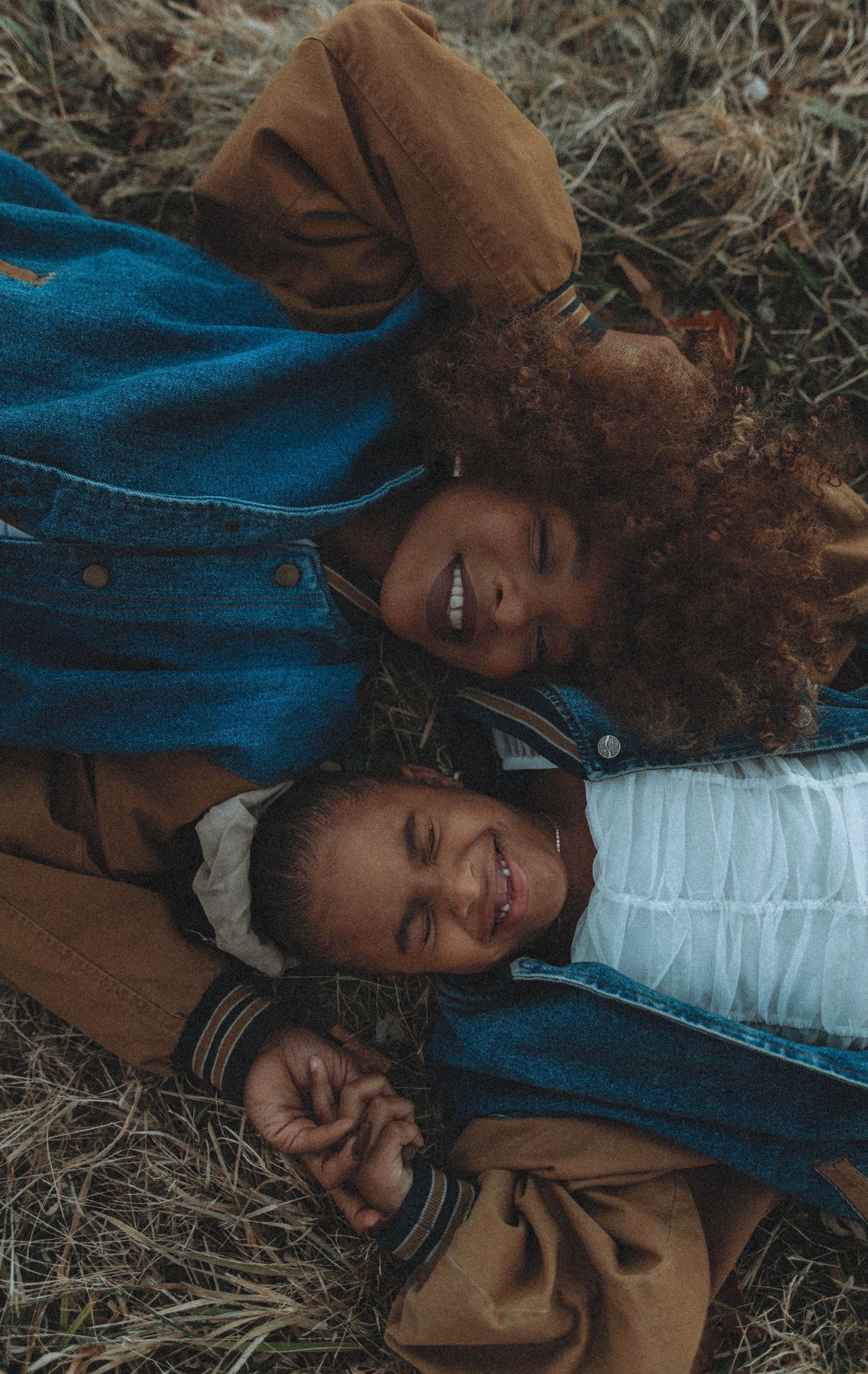 A mother and daughter share a joyful moment lying on grass, eyes closed, outdoors.