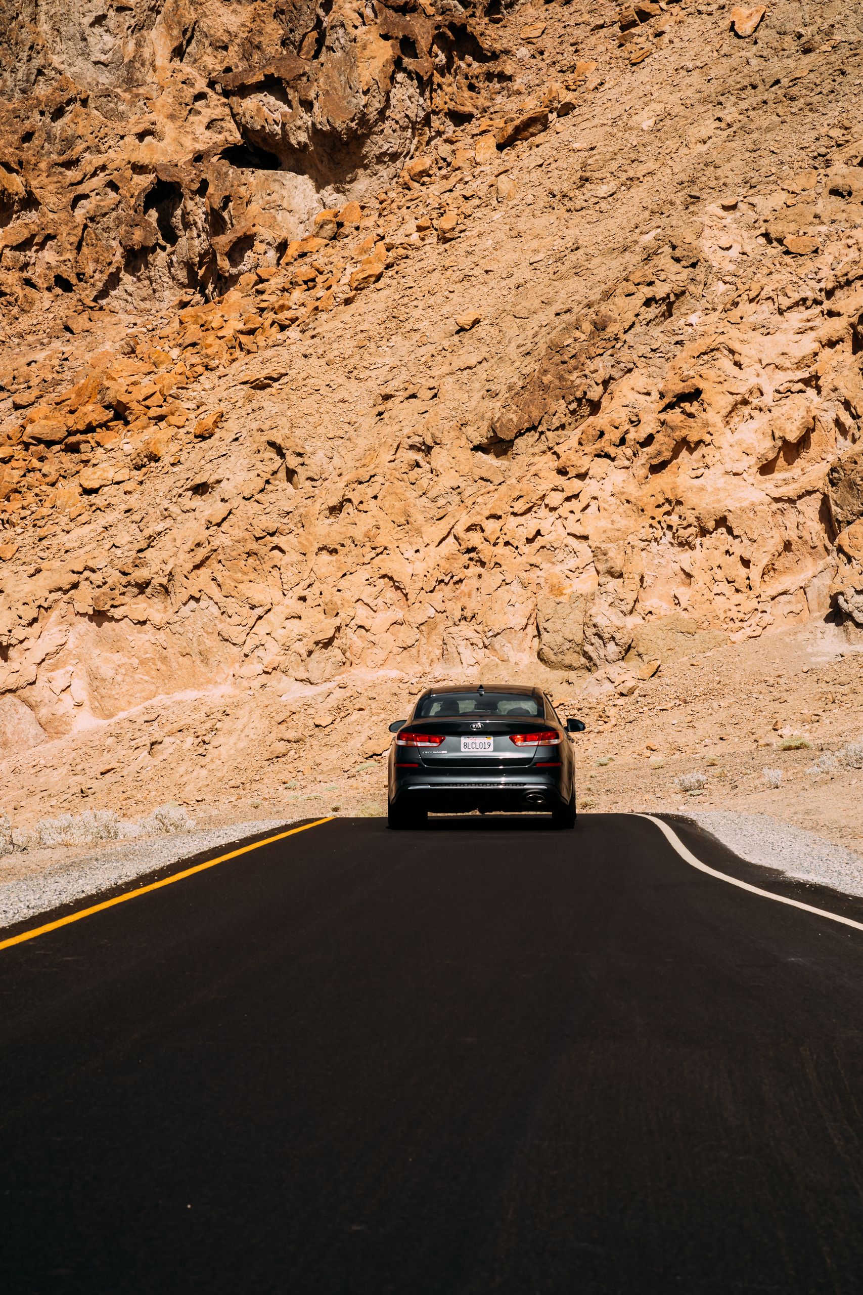 A solitary car driving on an asphalt road through arid desert rocks under daylight.