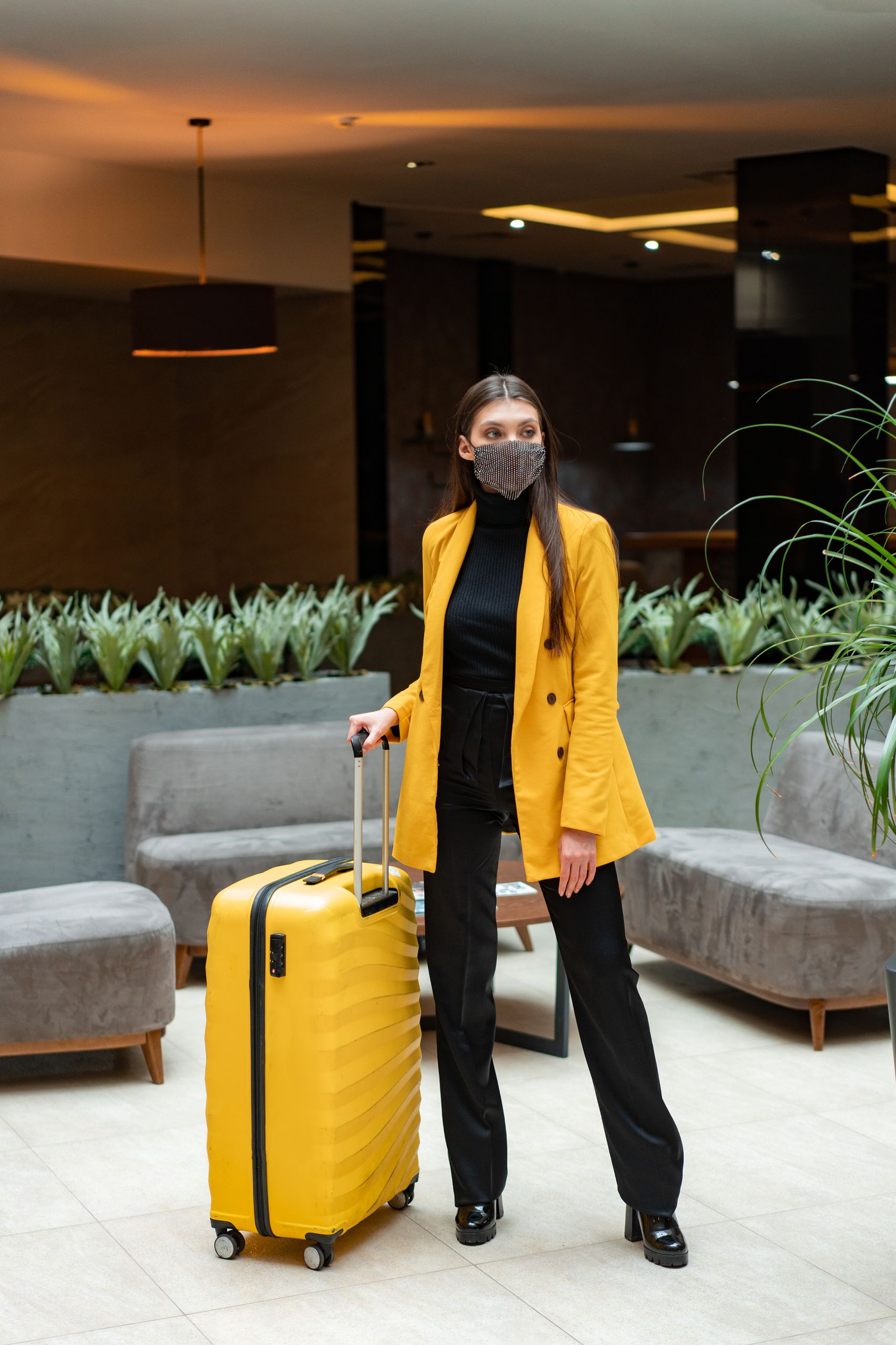 Stylish woman with yellow suitcase and face mask in a modern hotel lobby.