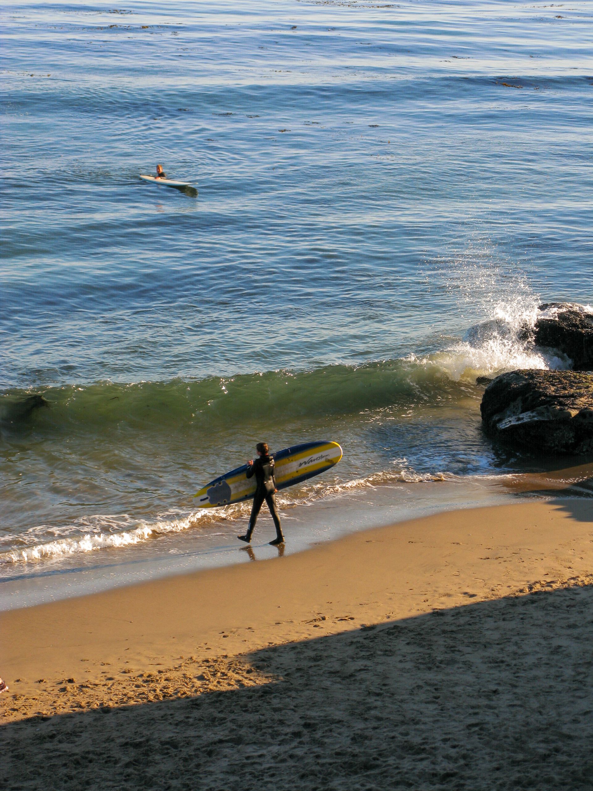 Surfer carrying board on sandy Santa Cruz beach with waves crashing in the background.