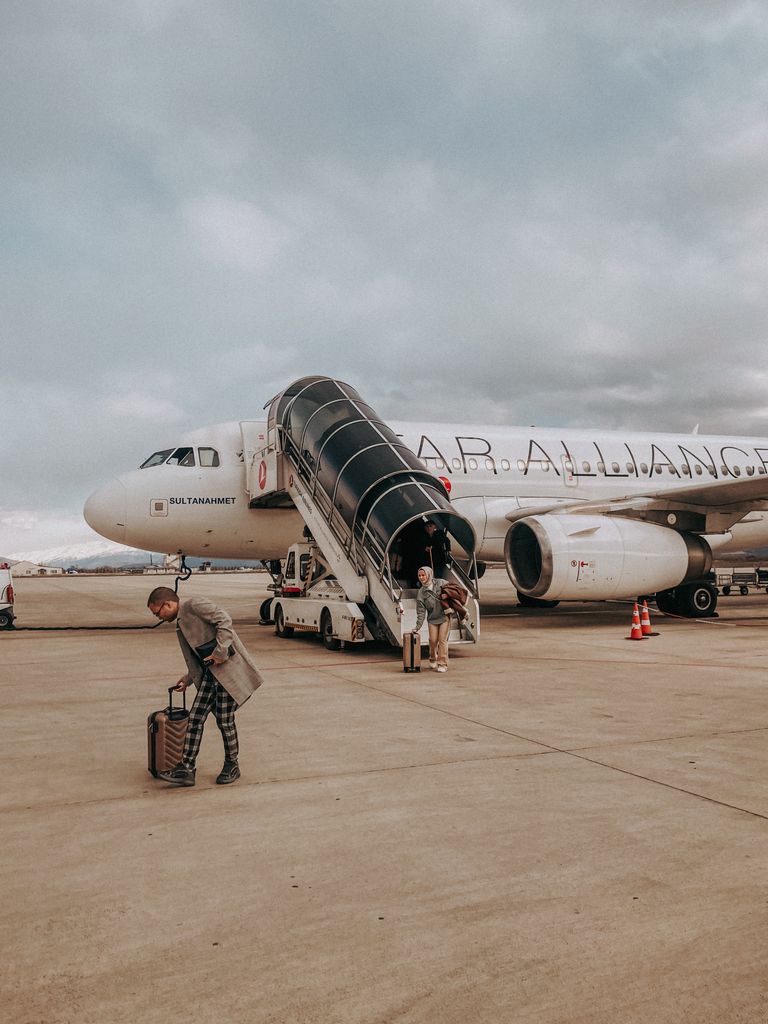 Travelers exiting aircraft onto tarmac via mobile stairs under cloudy sky.