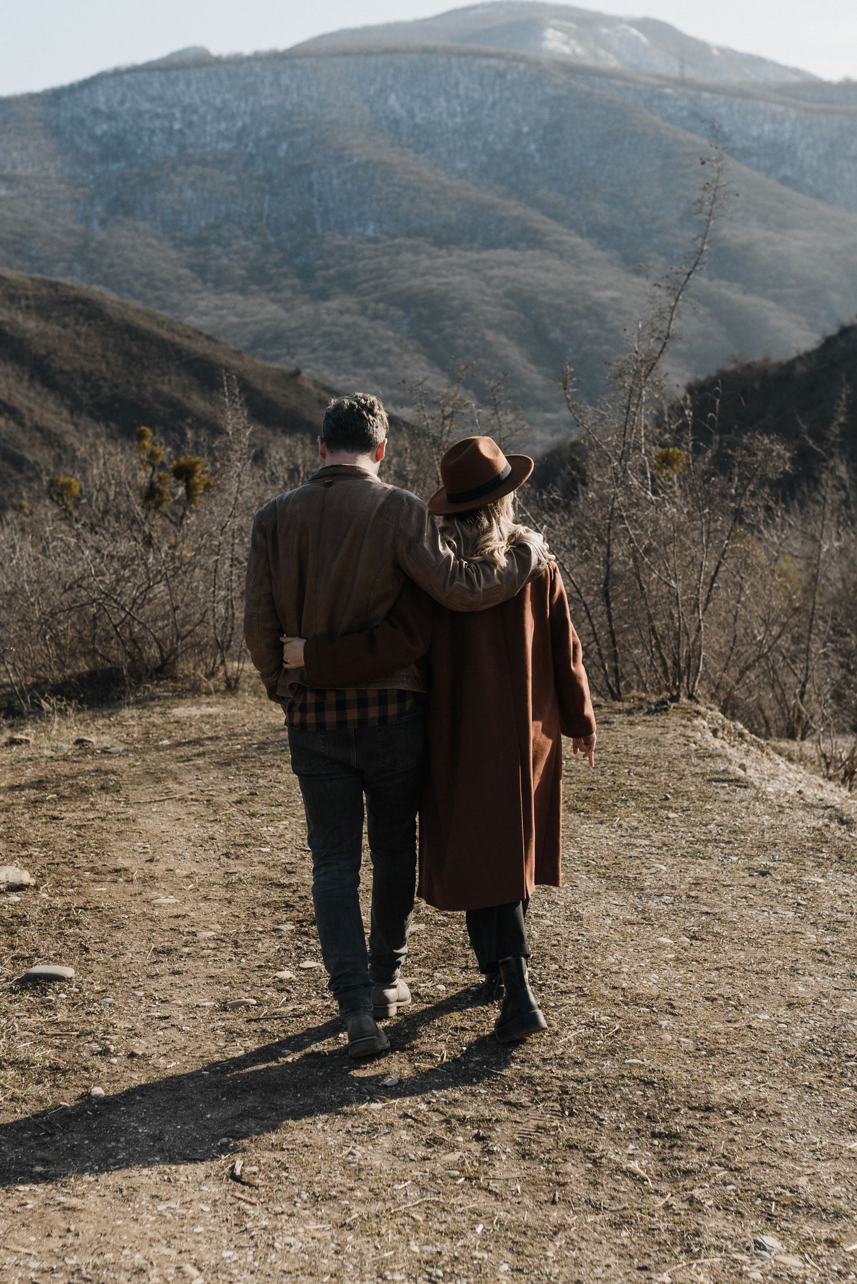 A couple in brown coats walking and embracing on a mountain trail with scenic views.