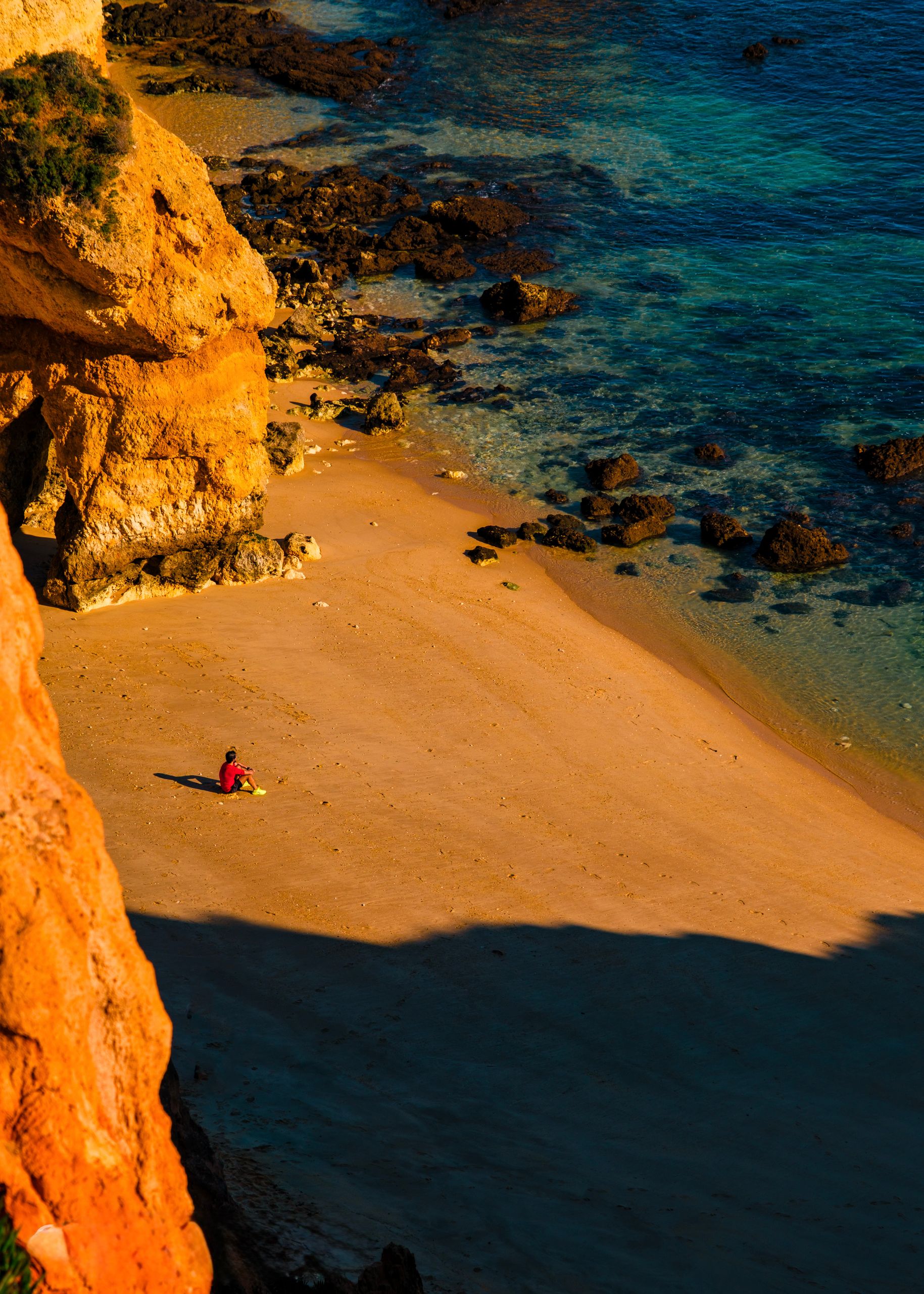 A lone person sits on a sandy beach at sunset beside rugged cliffs and blue ocean waters.