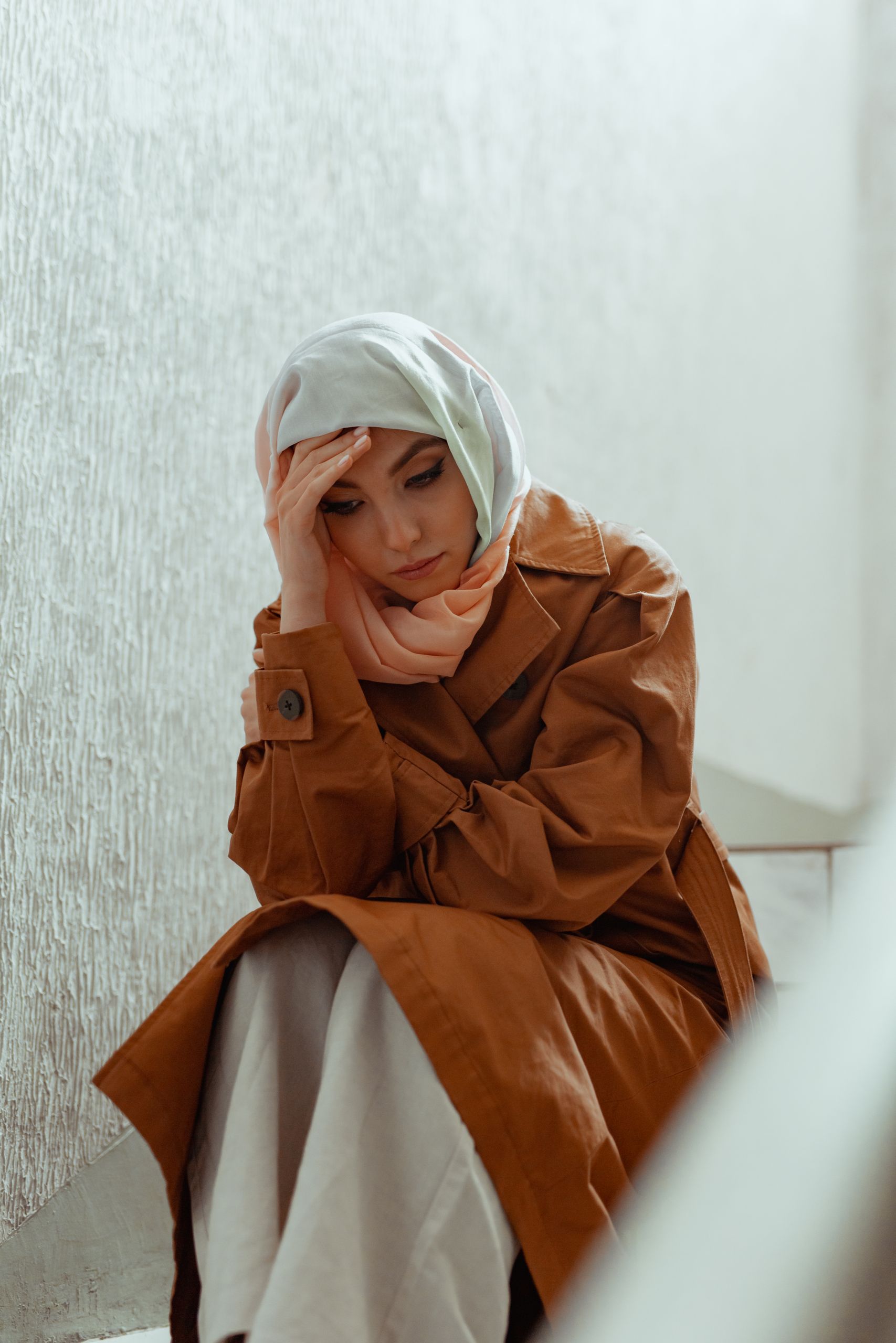 A woman in a hijab and brown coat sitting indoors, looking contemplative and thoughtful.