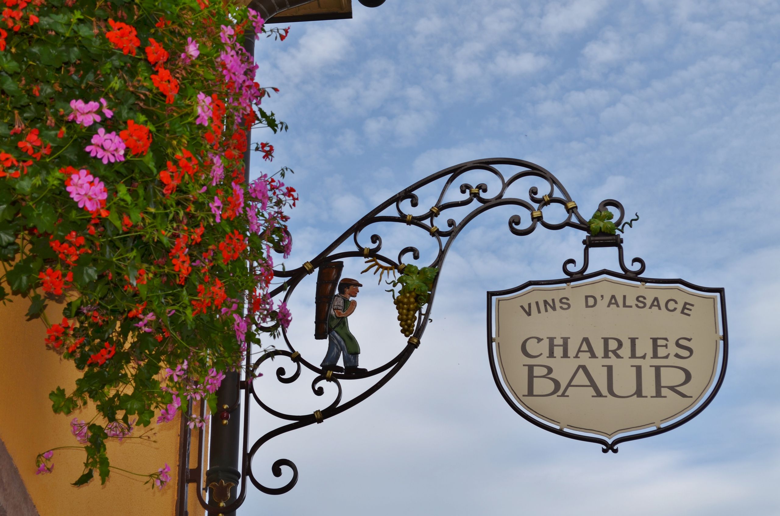 Ornate wine shop sign in Alsace, France with vibrant flowers and clear sky.