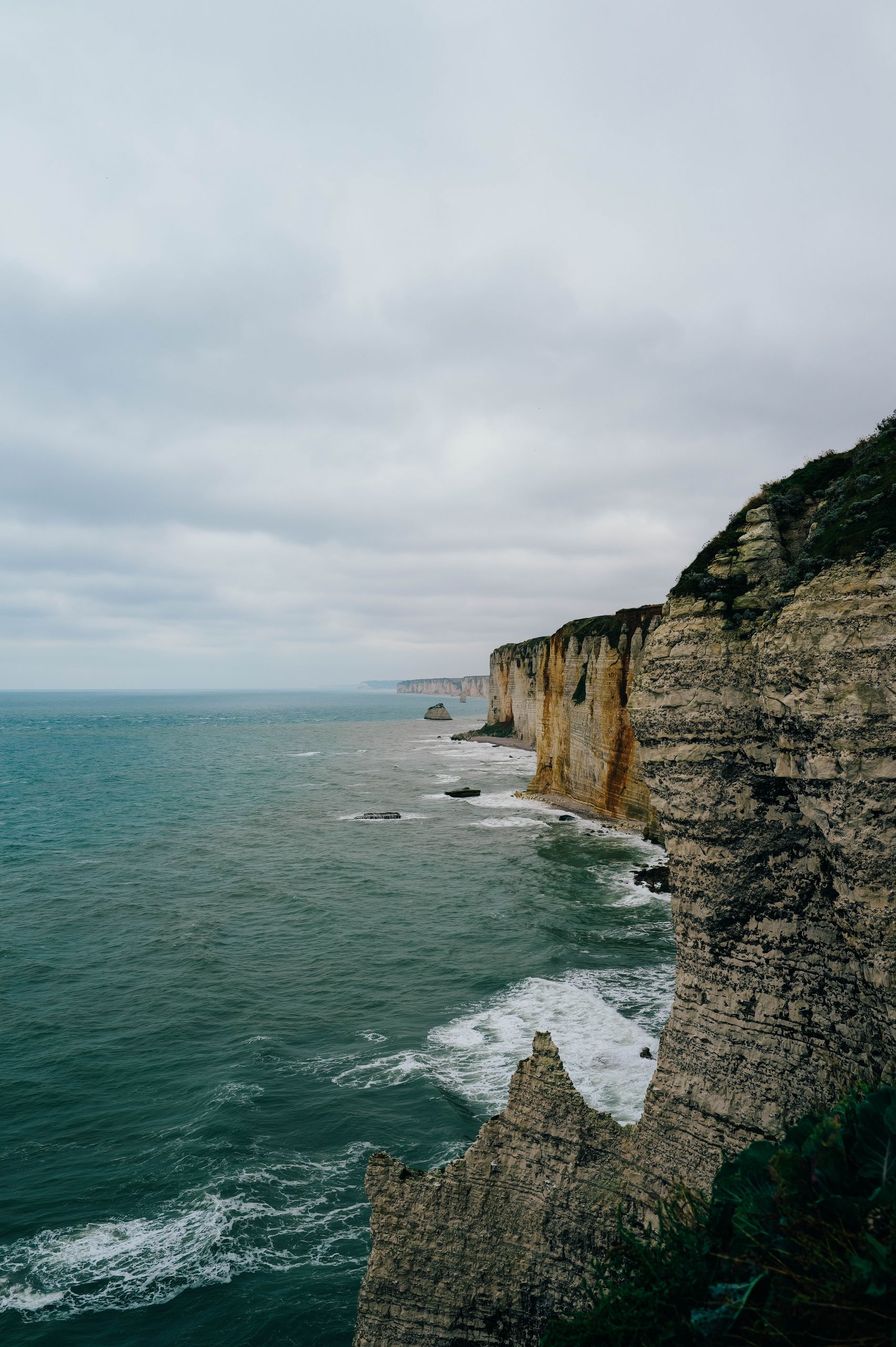 Panoramic view of Etretat's iconic cliffs with waves crashing below under a cloudy sky.