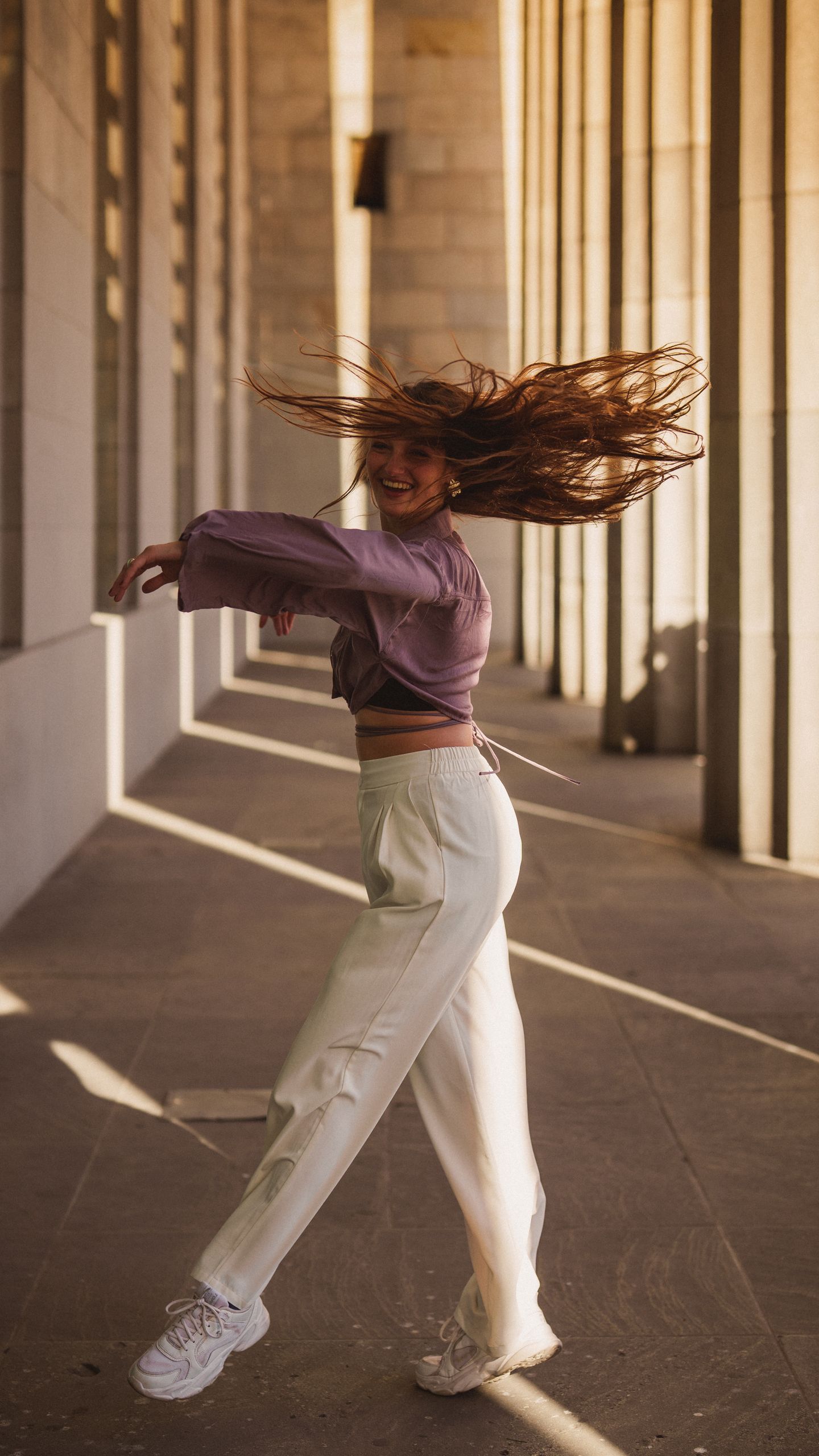 Young woman spins with joy in sunny outdoor corridor.